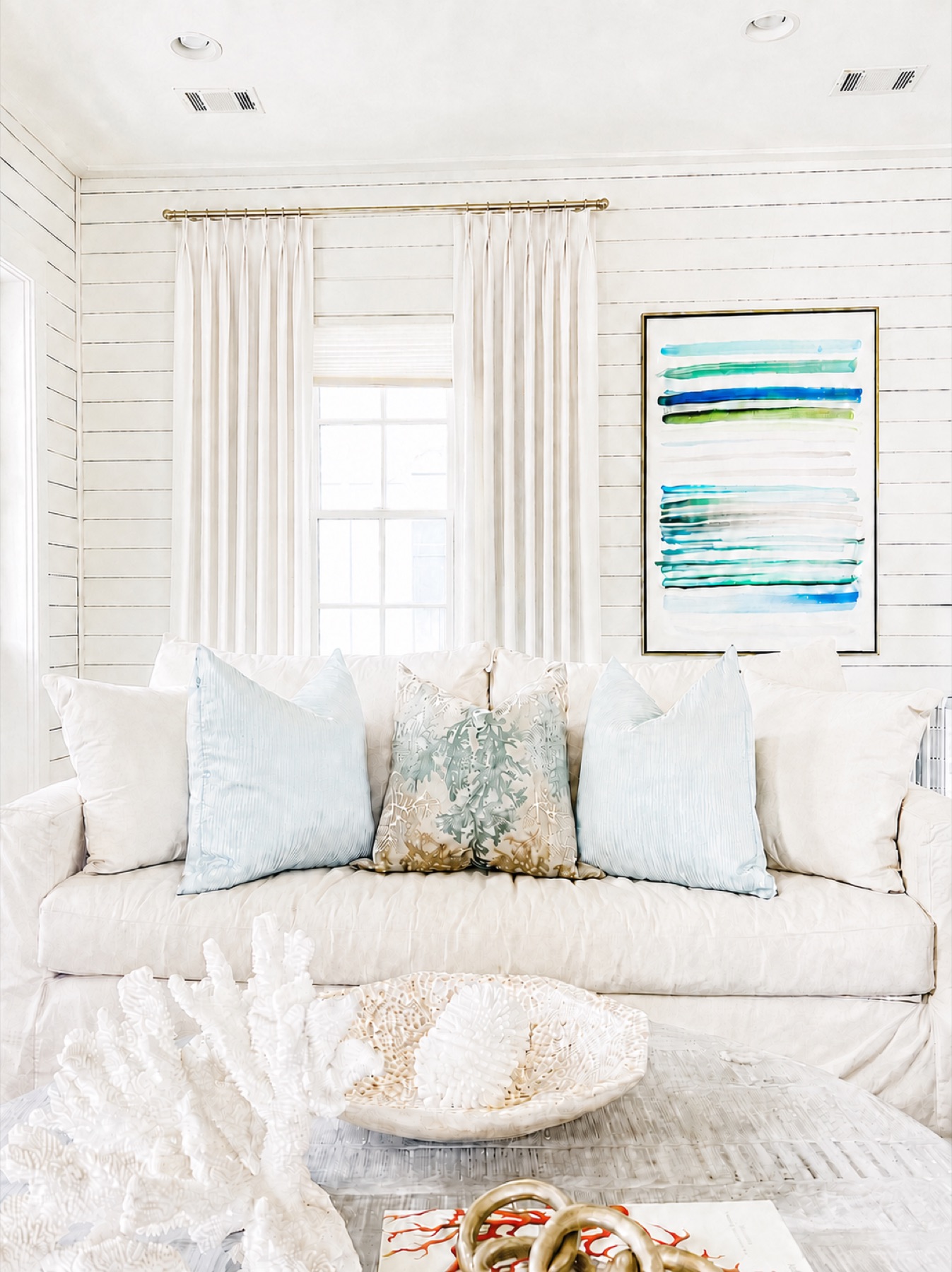 Living room with linen drapes on a brass rod, white shiplap walls, layered coastal pillows, and watercolor artwork